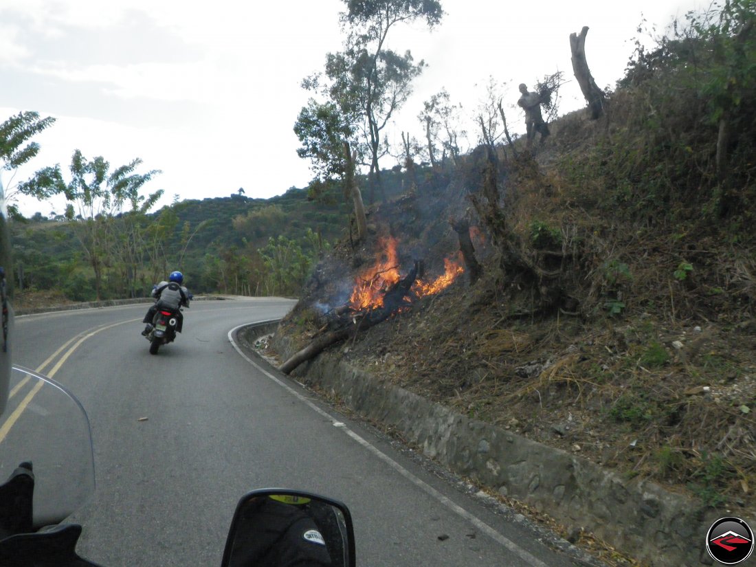 motorcycle riding around a corner past a burning tree on the side of the road