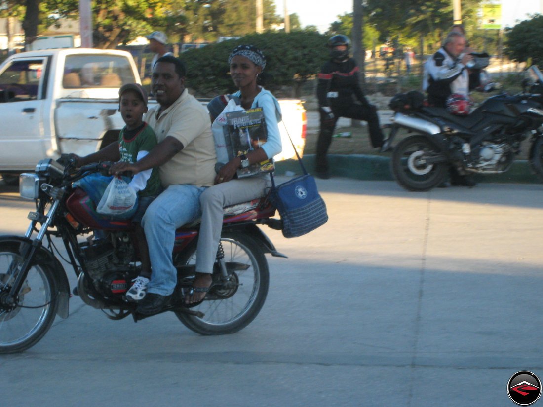 three people on a small motorcycle, mother, father and son, with mom carrying a toy transformer